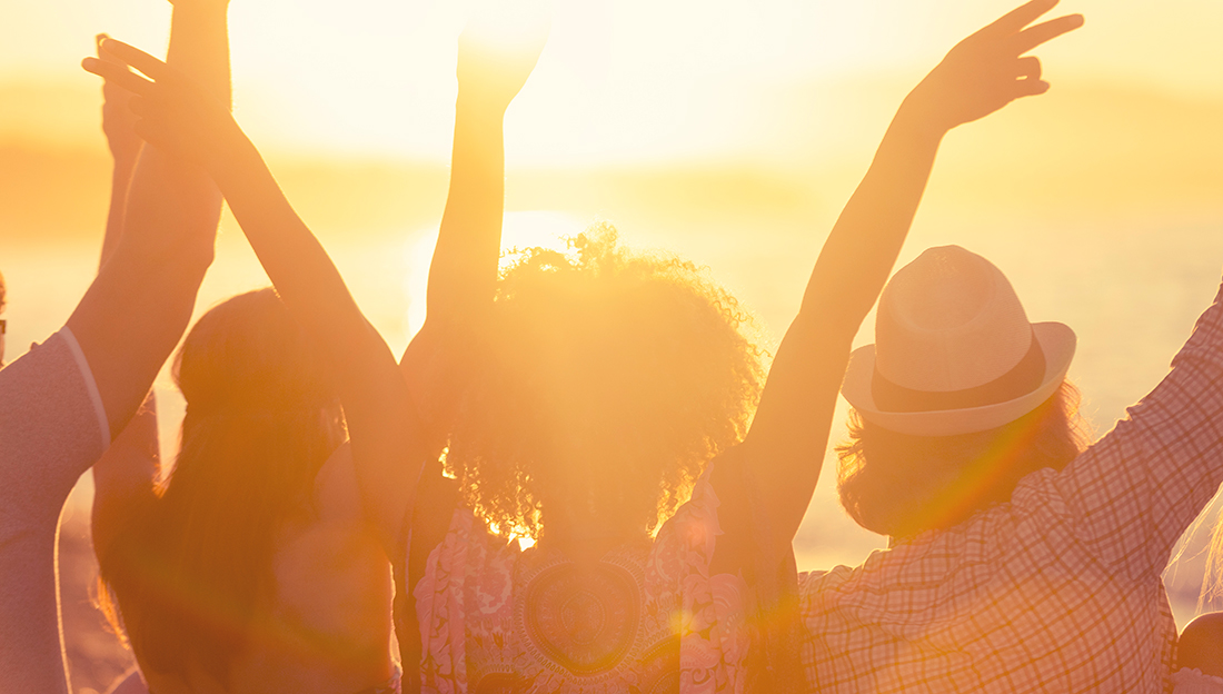 A group of people with raised arms celebrates the sunset on a beach. They enjoy the warm glow of the sun.