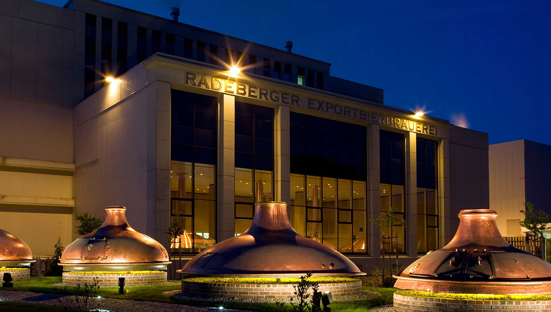A modern brewery building at night illuminated by spotlights with large copper kettles in front.