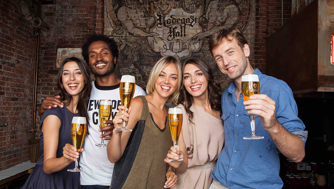 Five adults are smiling and holding glasses of beer at a bar with a rustic brick backdrop.